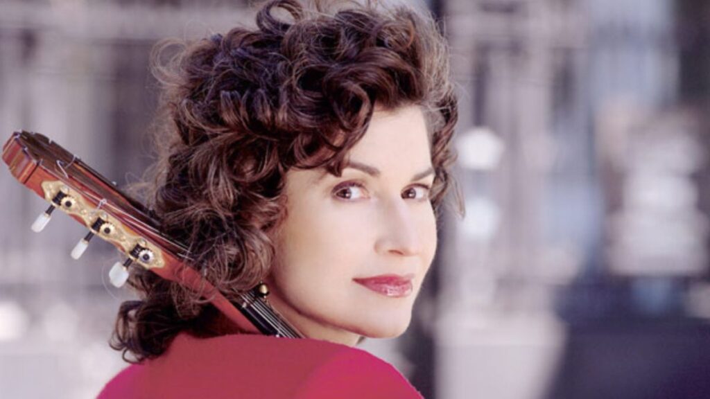 A woman with dark hair, holding a guitar and wearing a red sweater, looks over her shoulder at the camera.