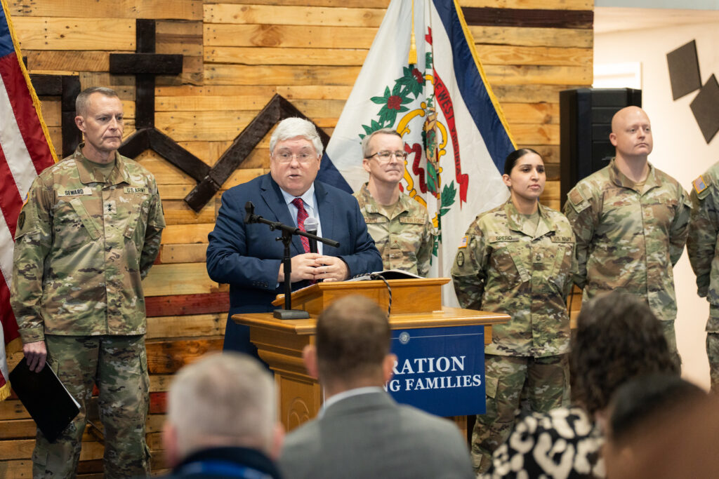 Man in suit stands in front of audience with national guard people on either side
