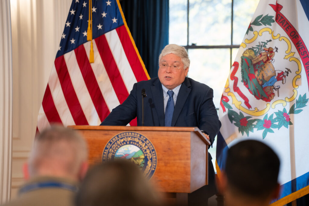 A man wearing a dark suit over a blue tie stands a lectern with a West Virginia seal on its front. Behind the man is a large window flanked by a U.S. flag and a West Virginia flag.