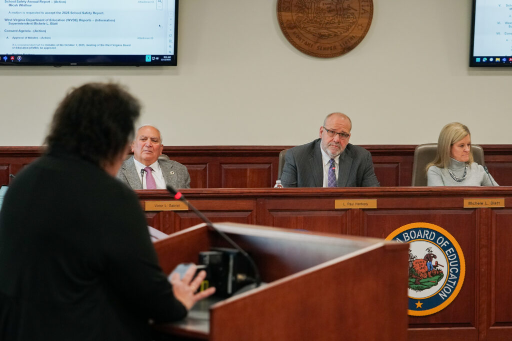 A person stands at a wooden lectern in the foreground. In main focus are three people two men wearing grey suits on the left and a woman on the right sit at a wooden dais below a wooden state seal.