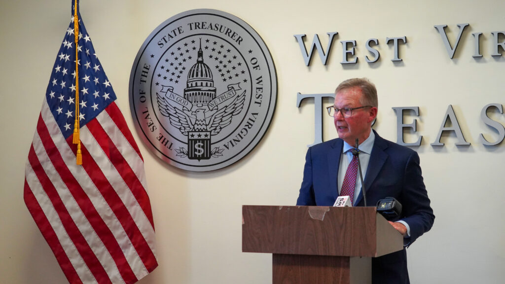 A man wearing glasses and a purple tie over a light blue shirt with a dark blue suit stands speaking at a wooden lectern. He stands in front of a wall that has partial lettering of "West Virginia Treasury", a seal of the Treasurer's office and an American flag.