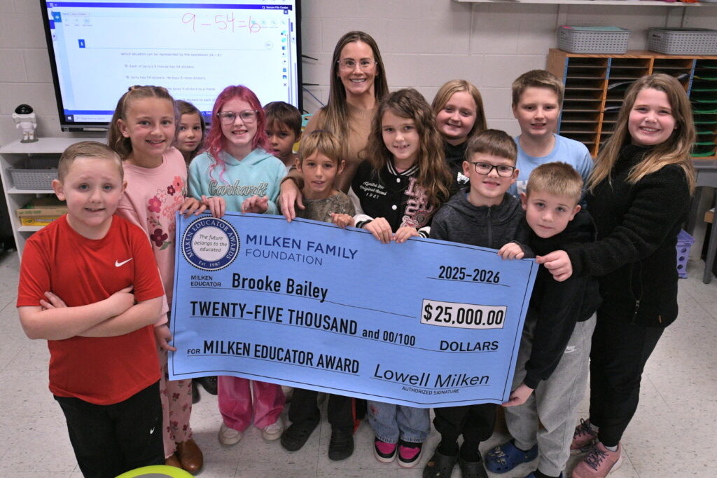 A group of children hold a blue novelty check for $20,000 made out to Brooke Bailey in a classroom in front of a projection screen. In the middle of the group stands a woman wearing glasses.