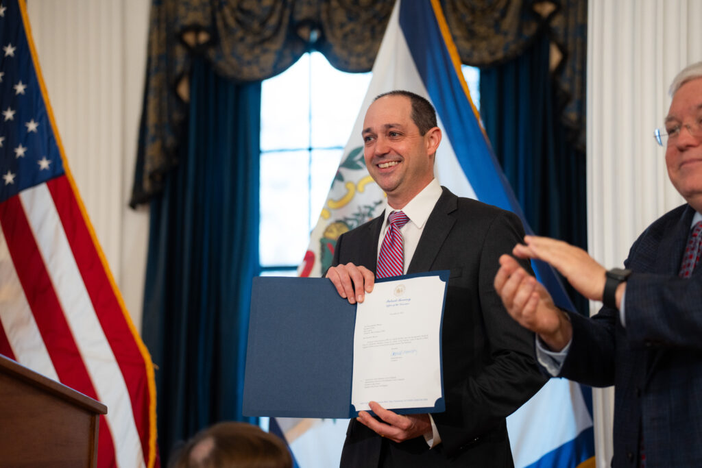 A man wearing a dark suit with a purple tie over a white dress shirt smiles as he holds a paper in a blue cover. He stands in front of a West Virginia flag in front of a large window. To his right can be seen a man clapping and to his left an American flag.