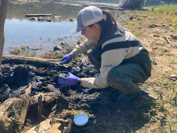 A woman crouching down at the bank of a river scooping black materials into a cup.  