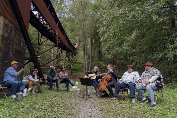 Eight people sitting in a semi-circle outside of a coal mining complex. Two people are playing instruments. 