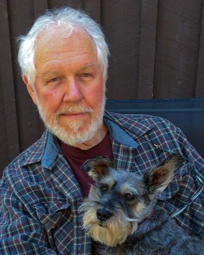 An elderly white man looks toward the camera. He holds a small dog in his lap.