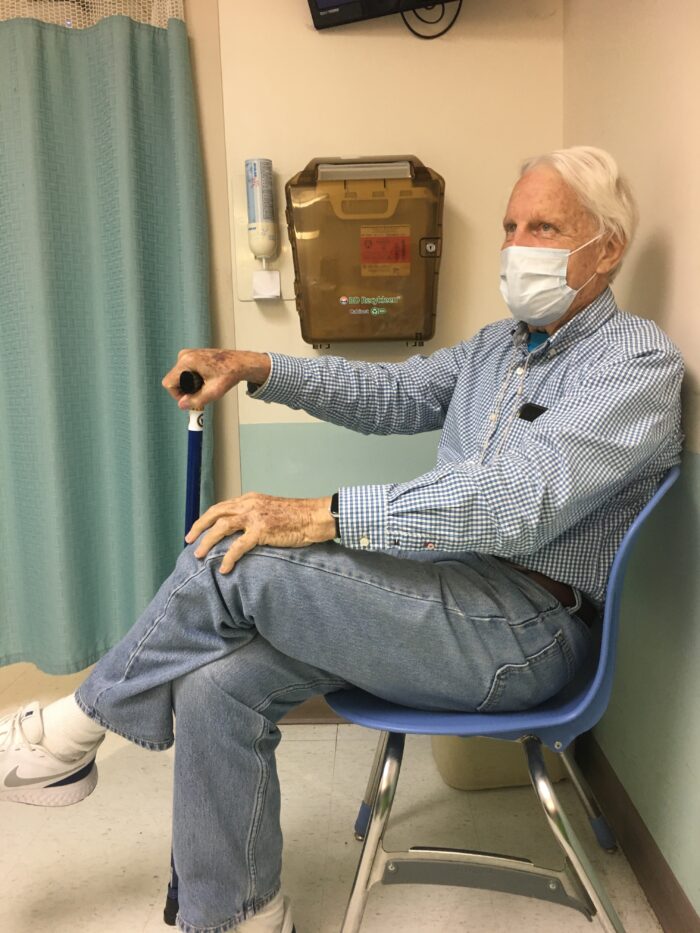 An elderly white man wearing a medical mask sits in a doctor's office. He holds a cane.