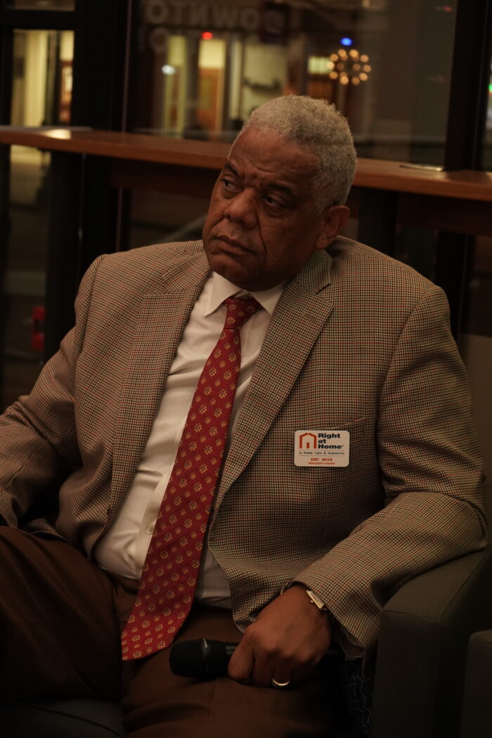 An older Black man looks off camera as if listening to another speaker. He is formally dressed and is holding a microphone while sitting on a stage.