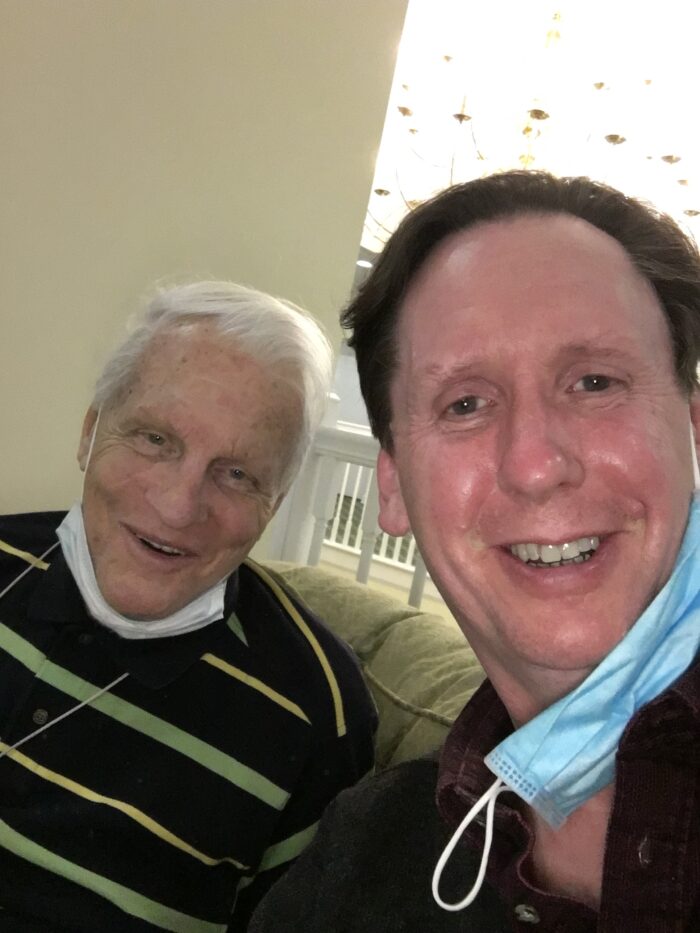 An elderly white man sits next to an adult man in his early 60s. They both smile for the camera in a selfie style.