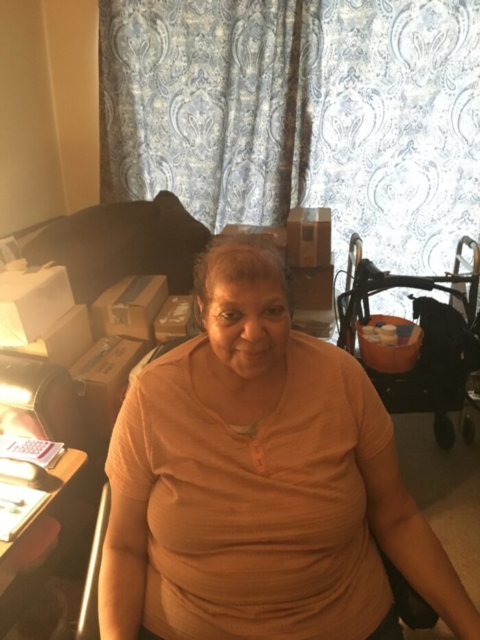 An elderly Black woman sits in a chair in her home and looks up at the camera.