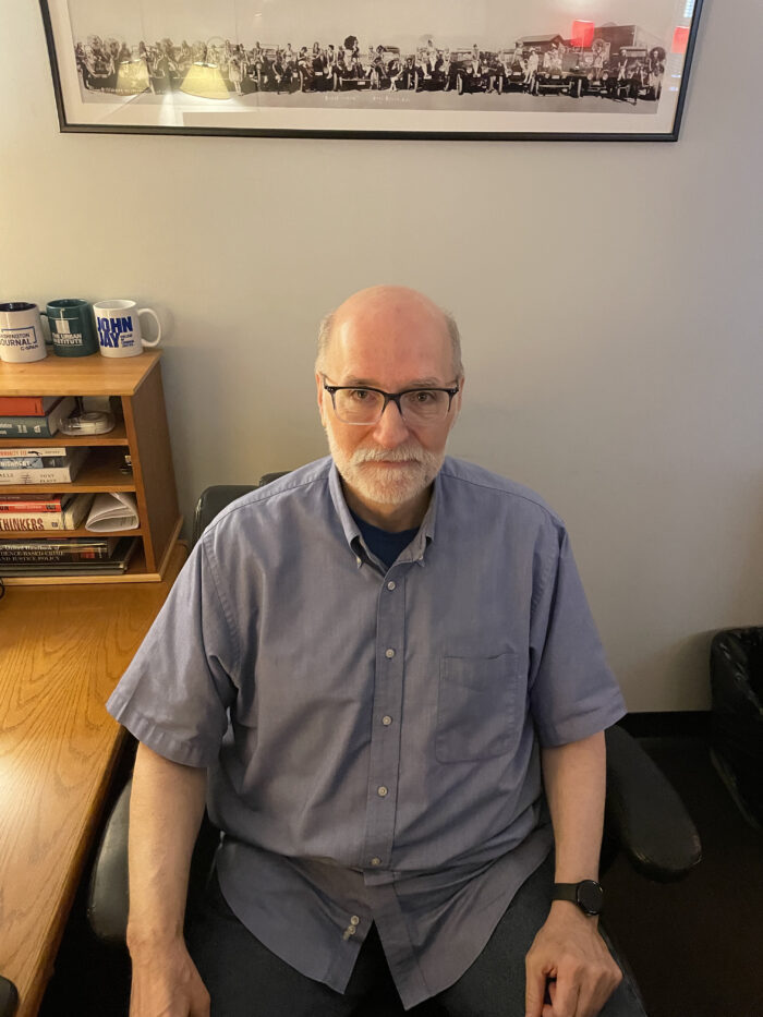 An older man sits in a desk chair and looks up toward the camera. He wears glasses, has a short white beard and wears a light blue button-up shirt.