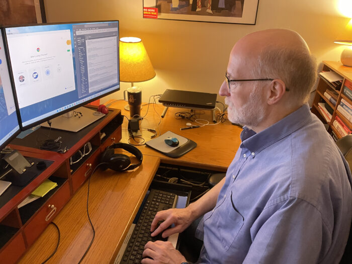 An older man faces toward two computer screens on a desk. He has a white beard, wears glasses and light blue button-up shirt.