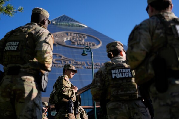 Four National Guard troops are shown from behind with the camera angled up from the ground. The troops stand in front of a building on a clear, sunny day.