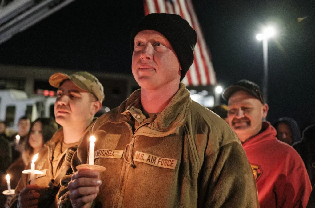 A Air National Guard member is shown at a candlelight vigil. He holds a small candle. Two other men are seen behind him with dozens more people seen in the background. An American flag is seen behind his head.
