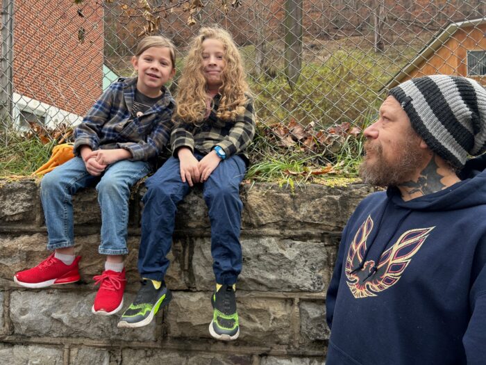 Two young boys sit on a stone wall and smile for the camera. Their father looks up at them as the photo is taken.
