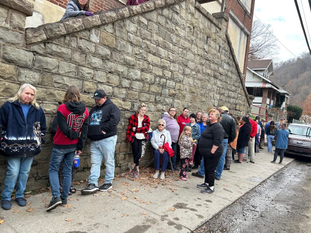 More than a dozen people stand in a line outside and lean against a stone wall.