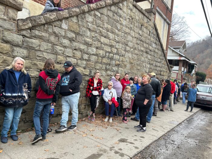 More than a dozen people stand in a line outside and lean against a stone wall.