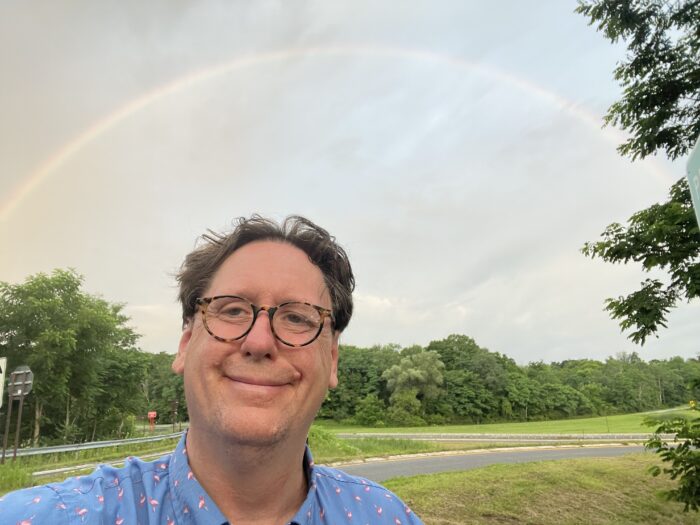 A man takes a selfie outside against the backdrop of a cloudy sky after a storm. There is a rainbow visible.
