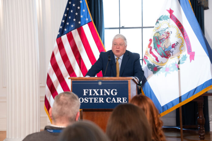 A man with white hair wears wire frame glasses and a gold tie over a light blue buttonup shirt and a navy blue suit. He stands at a wooden lectern with a sign affixed to the front that reads "Fixing Foster Care". He stands in front of a large window with an American flag and a West Virginia flag on either side.