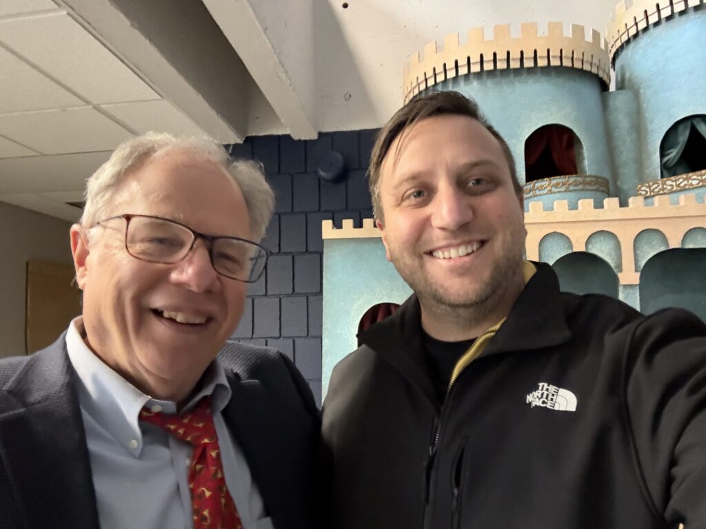 Two men, one older and one younger, stand next to each other in front of a TV set.