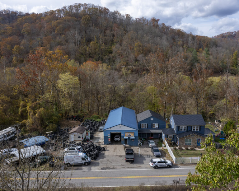 A service center photograph from faraway. The building is blue and is against a hill full of trees that have lost leaves due to fall weather changes.