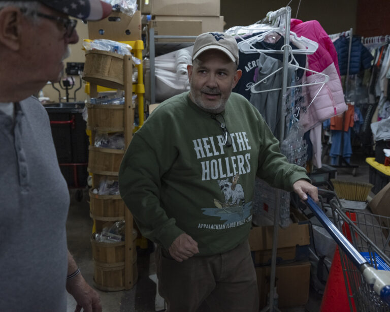 A man stands inside a food bank. He wears a green sweatshirt and ball cap.