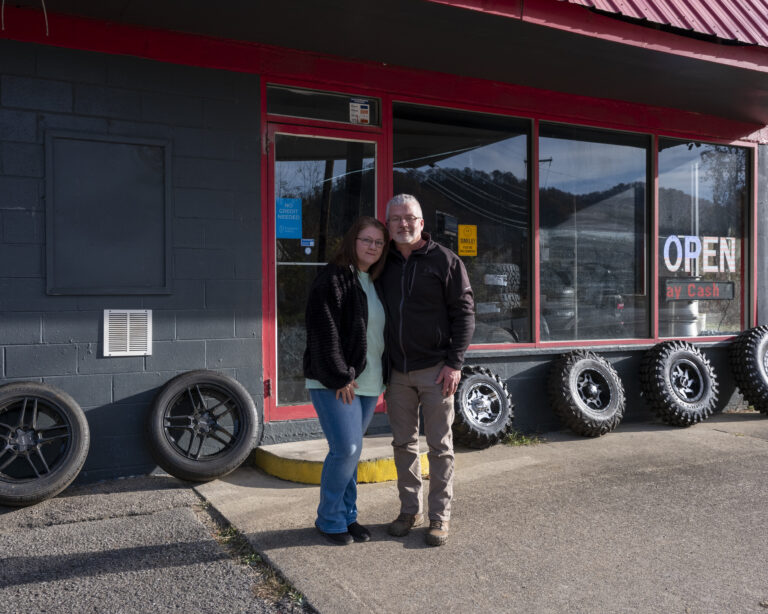 Two adults, a man and a woman, stand outside next to each other in front of a tire shop.