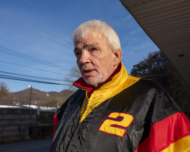 Close up photo of an older man with white hair wearing a black, yellow and red jacket.