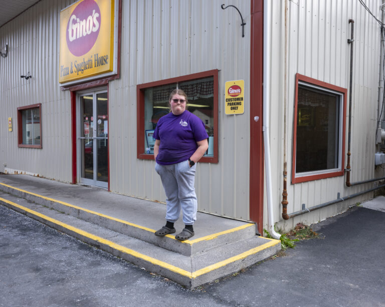 A woman stands on a street corner looking toward the camera. Behind her is a Gino's building and sign.