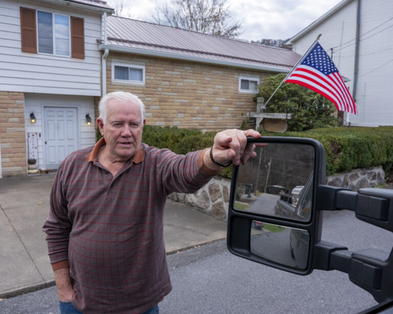 An older man stands next to the window of a truck on an overcast day. An American flag can be seen behind him hanging from the front porch of a house.