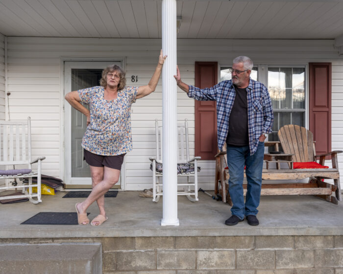 Two adults, a man and a woman, lean against a support beam on porch.