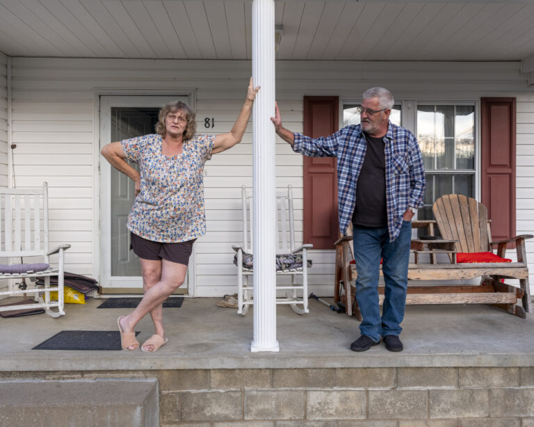 Two adults, a man and a woman, lean against a support beam on porch.