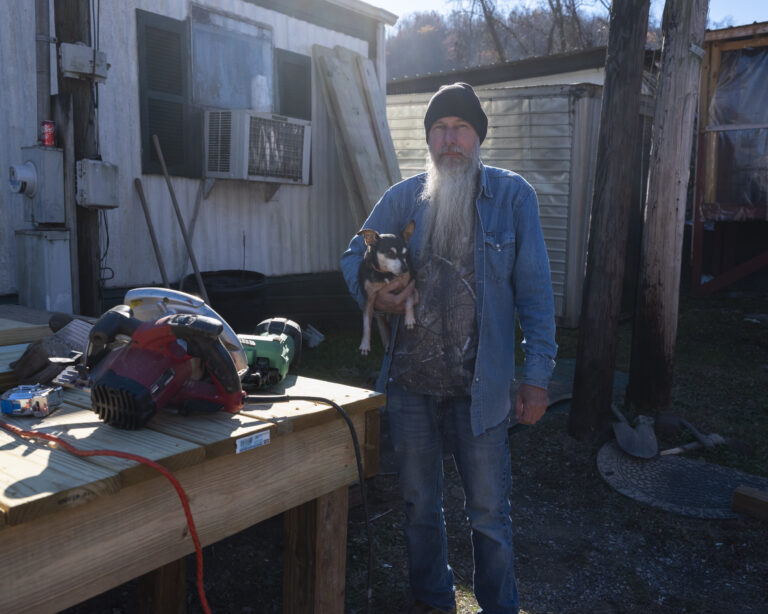 A man stands inside his home holding a small dog under his arm.