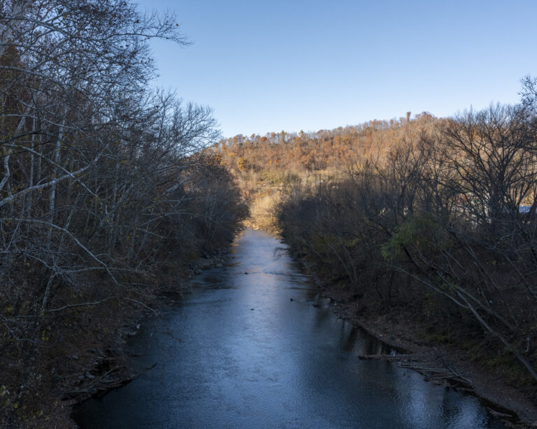 A photography of a river. Trees are seen on either side of the river bank. A hill at fall time is seen in the distance.