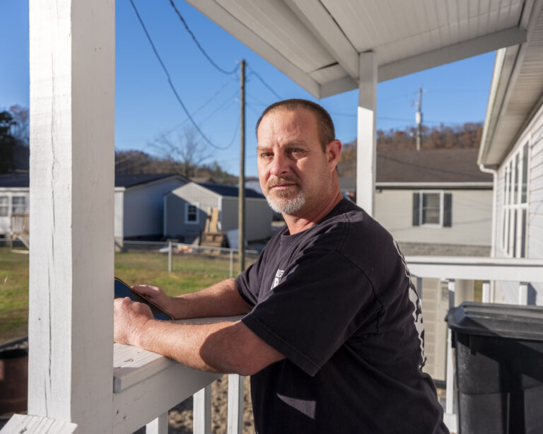 A man with graying hair leans against the railing of his porch on a sunny day.