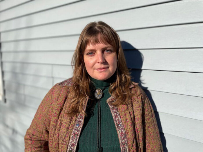 A white woman standing on the side of a building. She is wearing a green shirt and a brown jacket. 
