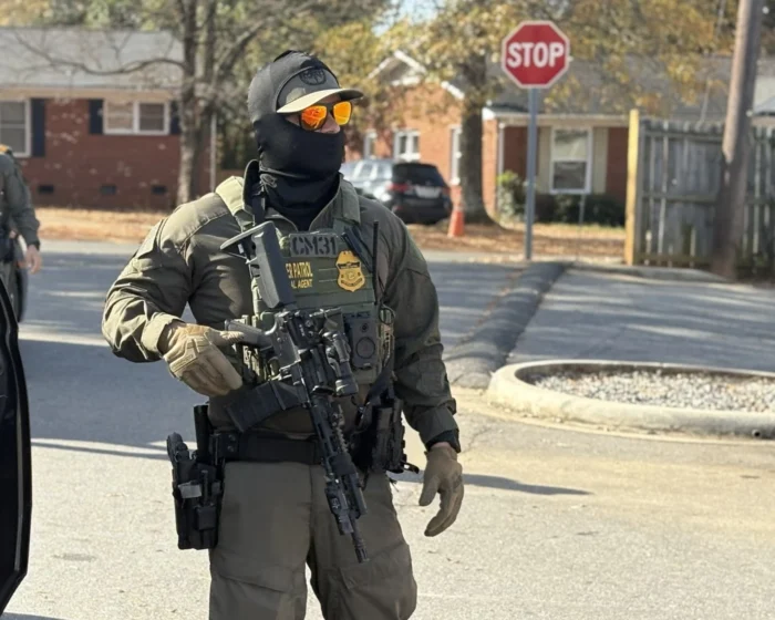 A man wearing military gear holding a gun. He is masked and standing in the middle of a street. 