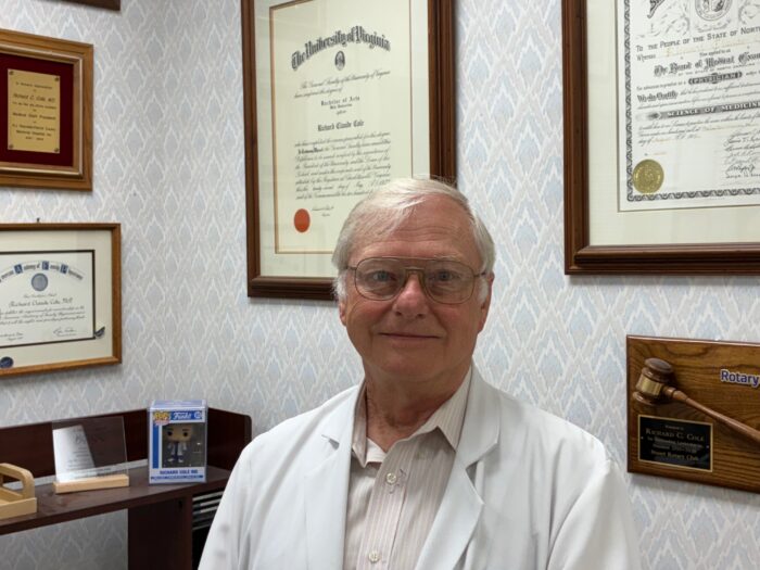 A white man sitting in his office. He is wearing a white doctor's coat. 