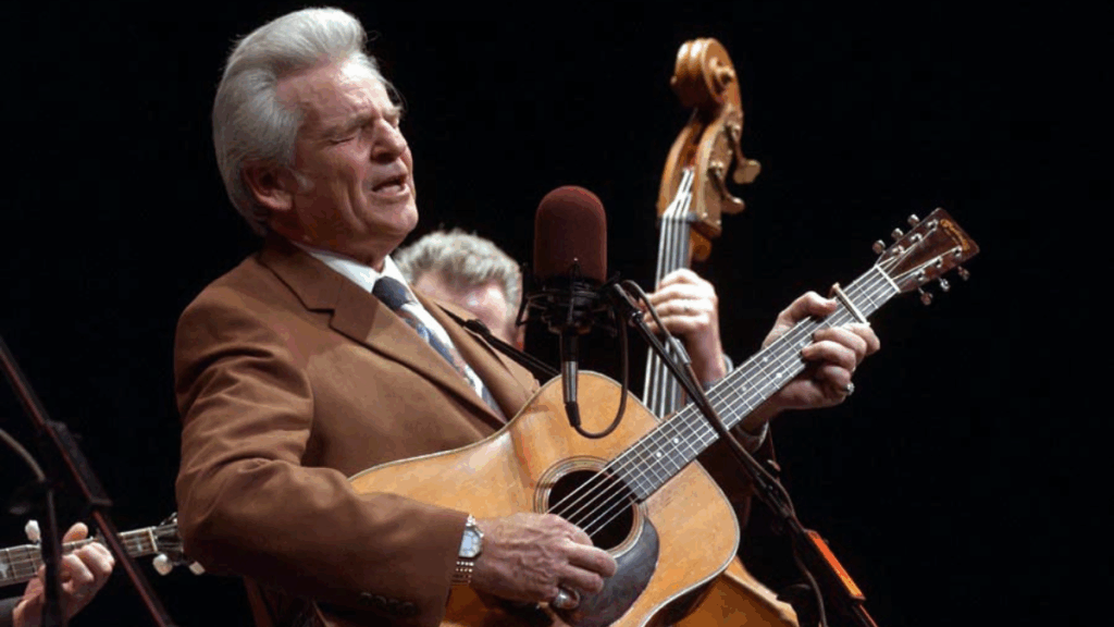 A man with white-gray hair sings into a microphone while playing a guitar. He wears a tan suit.