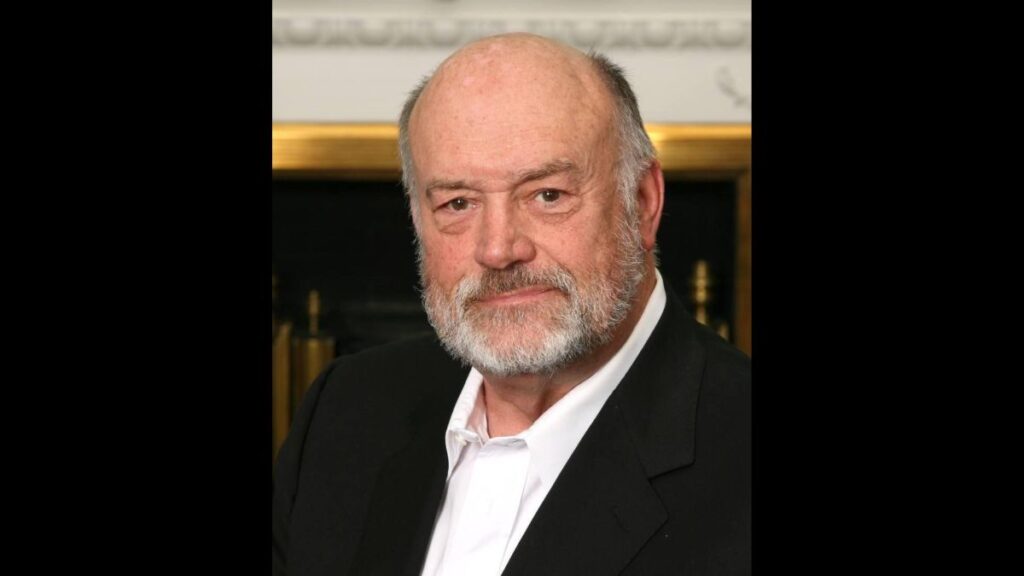 An older man with white, gray hair looks at the camera for a professional head shot. He wears a black suit jacket and a white button up shirt.