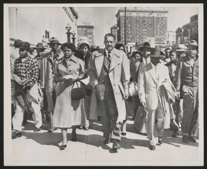 A black and white photo of Thurgood Marshall walks down a street as dozens of other people walk behind him.