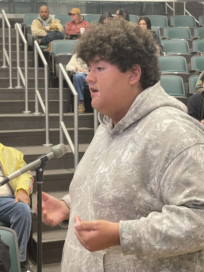 A young man with curly brown hairs speaks at a stand microphone toward panelists.