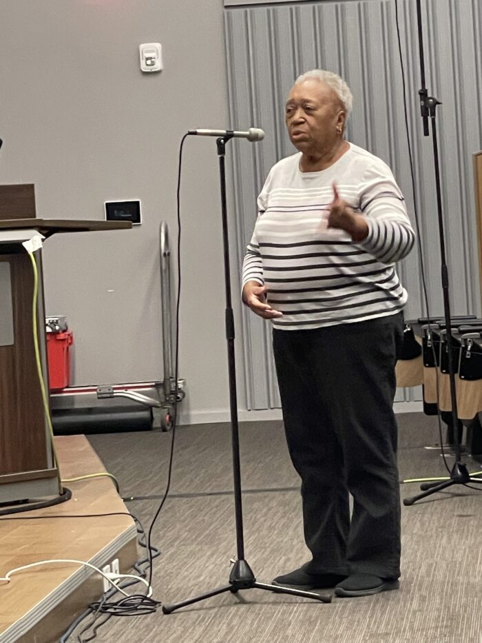 An older Black woman stands at a microphone and speaks to a panel.