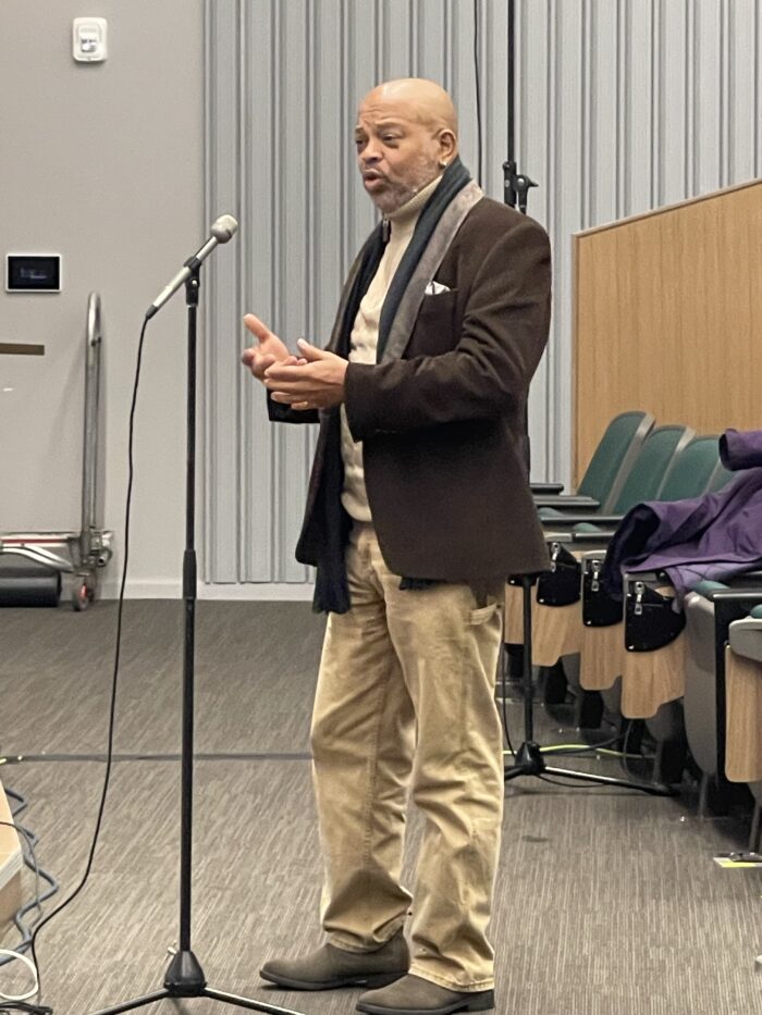An older Black man with a graying beard speaks at a stand microphone toward panelists.