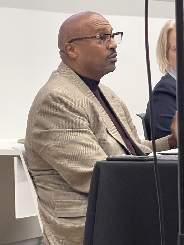 A close up photo of a Black man speaking from a panelists' table. He wears glasses and a tan suit jacket.