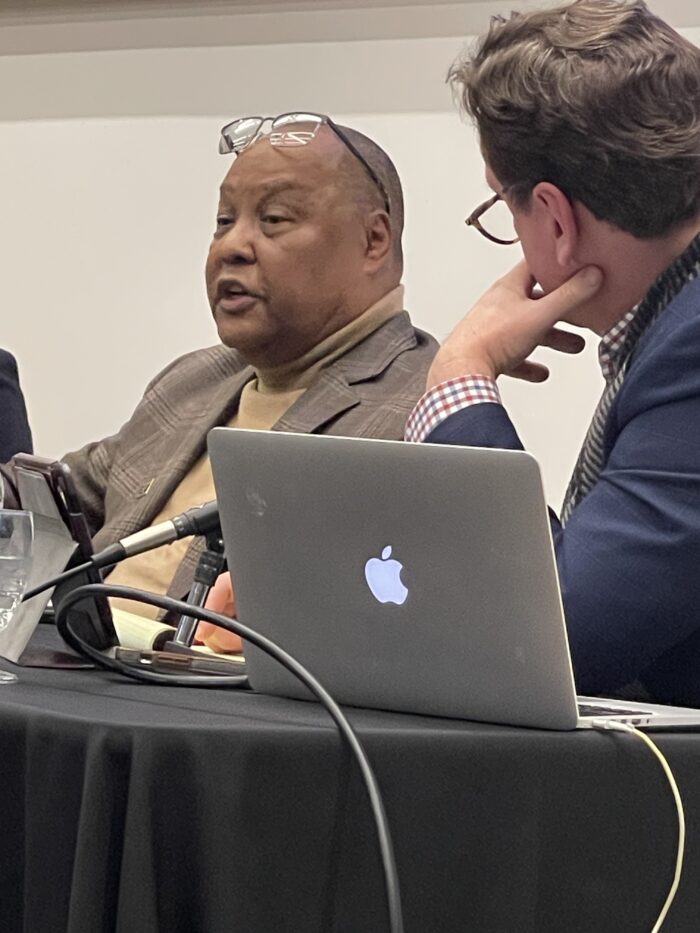 A Black man with glasses pushed up on his head addresses the audience from a panelist table. A white man with brown hair and glasses leans in to listen.