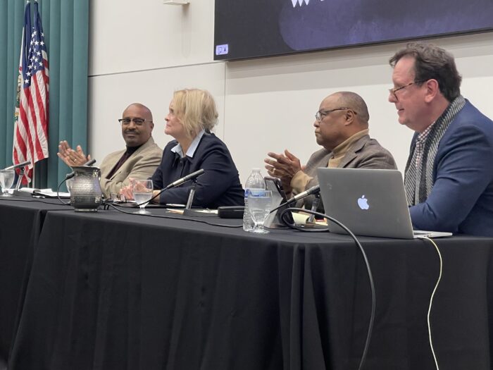 Four adults, three men and one woman, are seated at a table for a panel discussion. Two of the men are Black, while the woman and the third man are white.