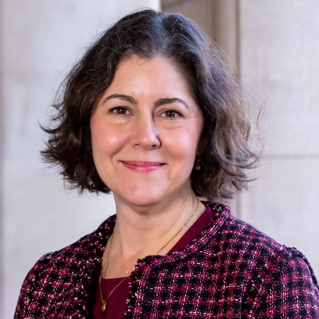 A woman wearing a hatch pattered red-purple and black jacket over a red-purple blouse stands facing the camera in front of an arcade of concrete columns