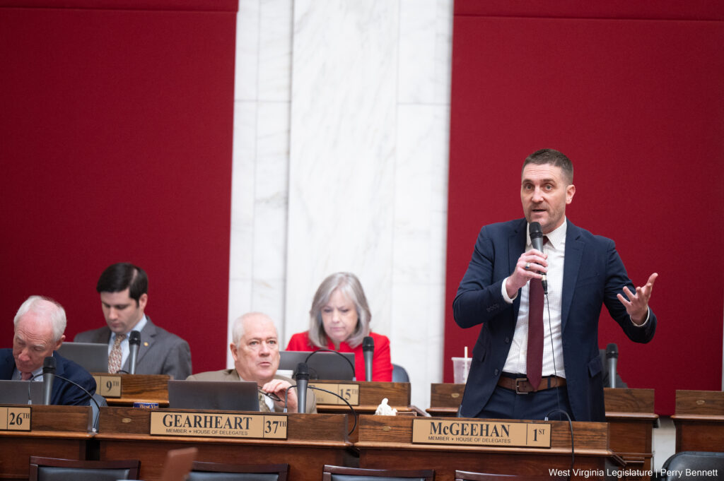 A man wearing a dark blue suit over a white button-up and a maroon tie stands behind a wooden desk with a bronze nameplate that reads "McGeehan 1st." He holds a wired microphone in his right hand while gesturing with his left. Four people are seated at similar desks to his right with their own bronze nameplates with black text. The man stands in front of a large scarlet wall panel over white marble.