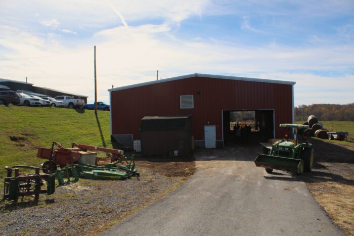 A red building with a bay opening has farm equipment arrayed outside of it, including a tractor. To the side hay bales are piled high. The building sits under a blue sky with wispy clouds. To the left is a ridge with cars parked above, and to the right is an open field with a treeline in fall colors in the far background.